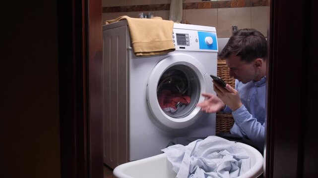 Young man experiencing anger and stress while doing laundry, yelling at his phone next to a broken washing machine with clothes inside, highlighting household chore frustrations