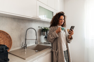 Woman in Robe Drinking Coffee at Home Kitchen While Checking Phone