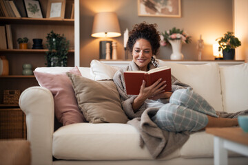 Fototapeta premium Cozy Woman Reading a Book on a Sofa in a Warm Living Room With Soft Lighting
