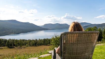 A woman sits on a chair in a garden, watching Gjevillvatnet lake surrounded by mountains in Oppdal Norway. She enjoys her free time in the summer sun and fresh air