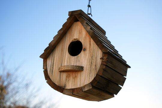 A rustic wooden birdhouse with a unique rounded bottom and shingled roof hangs against a clear, bright blue sky. The natural texture of the wood makes this ideal for themes of nature or wildlife.