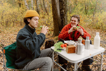 Two friends sit at a folding table in a vibrant autumn forest, enjoying homemade sandwiches. Golden...