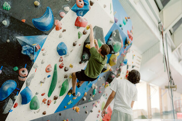 A young climber scales a colorful bouldering wall, showing determination and skill. A coach observes closely, ready to offer support and encouragement in a vibrant indoor climbing environment. © m-art
