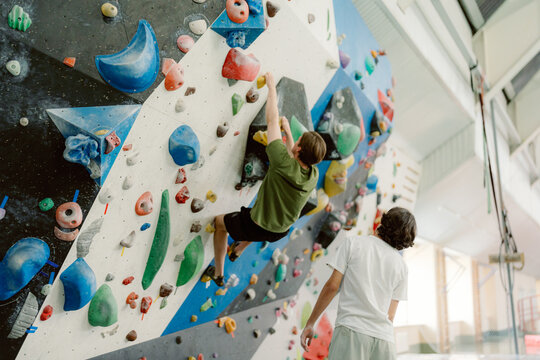 A young climber scales a colorful bouldering wall, showing determination and skill. A coach observes closely, ready to offer support and encouragement in a vibrant indoor climbing environment.