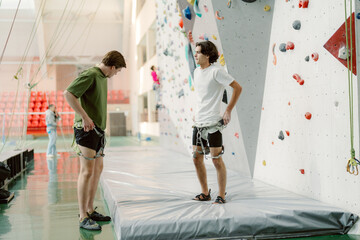 Two young men get ready for their climbing adventure at an indoor rock climbing gym. They check their harnesses and safety gear while surrounded by colorful climbing walls.