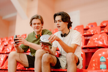 Two young men sit on red stadium seats, sharing their love for music. They eagerly hold a portable...