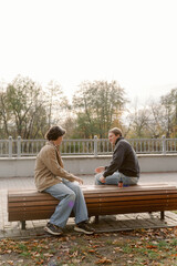 Fototapeta premium Two friends sit on a wooden bench in a peaceful park during late afternoon. Trees and soft clouds create a serene backdrop as they engage in cheerful conversation surrounded by nature.