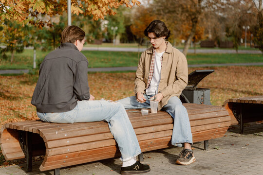 Two young men sit on a wooden bench in a park surrounded by vibrant autumn leaves. They are engaged in conversation, sharing drinks and laughter under the warm sun.