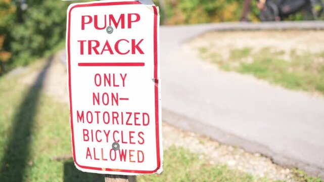 pump track sign along empty curving path early morning sunlight casting long shadows, red and white sign reads only non-motorized bicycles allowed, smooth asphalt track, green trees, calm