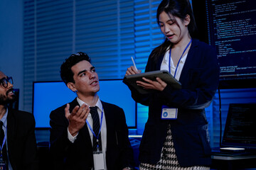 Software engineers and data scientists analyze AI code. An Asian female programmer holds a tablet while her Caucasian and Indian male colleagues discuss the system.
