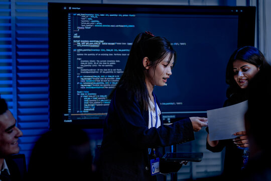An Indian female developer leads a code review, holding a report. She explains a Python script for an AI or cybersecurity system to her team in a dark SOC Security Operations Center - Powered by Adobe