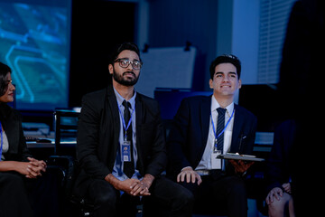 A large IT department meeting in a command center (SOC). An Indian analyst holds a tablet, listening with his team to a cybersecurity update on an AI system.