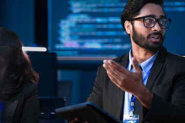 n Indian businessman with glasses discusses a project with his smiling Caucasian male colleague. They are having a positive team discussion in a modern, high-tech office.