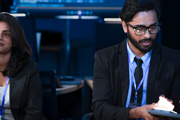 An Indian businessman holds a tablet while his Caucasian colleague uses it. The diverse team is collaborating on a project in a modern tech office at night.