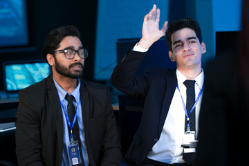 An Indian developer holds a tablet while a Caucasian engineer interacts with the screen. They are analyzing AI data or a cybersecurity system in a SOC.
