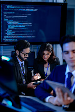 An AI research team brainstorms late night. Innovators discuss machine learning model architecture using a tablet while reviewing training data code displayed on a large screen.