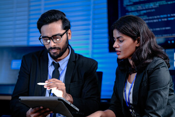 Code deep dive briefing. Experienced male developer explains software architecture or complex algorithm to female engineer with code visible on screen in background tech hub.