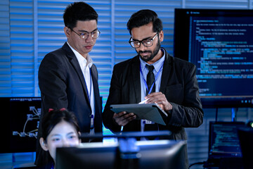 Diverse male professionals strategize late night modern tech hub focused review digital tablet stylus discussing project data code screens intense collaboration synergy achieved.