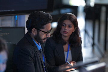 Focused diverse professionals collaborate late night in a modern tech office. Colleagues discuss project details using a tablet with stylus while code runs on a large screen.