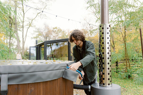 A young man adjusts the cover of a hot tub in a lush backyard during autumn. The scene showcases colorful leaves and a cozy atmosphere created by nearby trees and modern architecture.