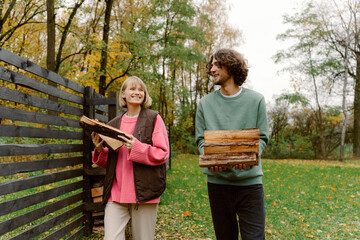 Naklejka premium In a tranquil park during fall, a couple smiles as they carry firewood together. Bright autumn leaves surround them, creating a warm and cozy atmosphere filled with laughter.