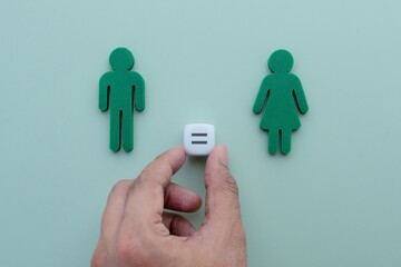 A hand places a white die with an equal sign between green male and female figures. Gender equality, fair rights, balance, and equal opportunity concept.