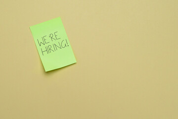 A green sticky note with the handwritten message WE'RE HIRING! on a plain background. Job openings, recruitment, talent acquisition, and employment concept. Copy space for text.