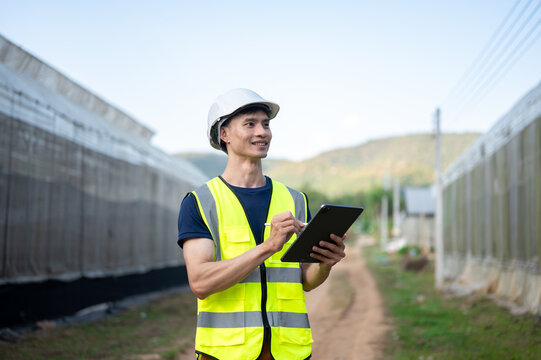 Asian engineer man wearing safety vest and hard hat helmet write in tablet standing aside greenhouse