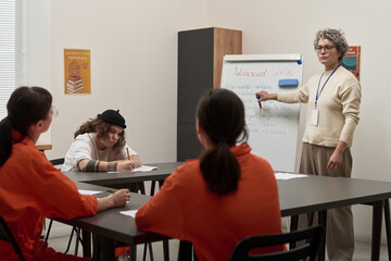 Obraz premium Middle aged Caucasian woman teaching English grammar to group of young adult women in orange prison uniforms, students sitting at tables writing notes during prison education class