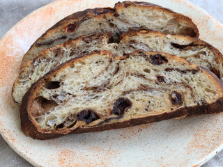 Artisan sourdough bread with chocolate and cherry slices on handmade ceramic plate, close-up of natural yeast loaf with swirled crumb texture and dried fruit pieces. The naturally leavened loaf