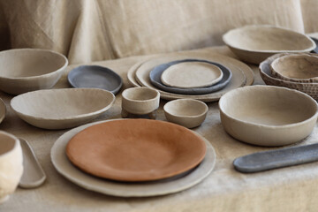 Assortment of handmade clay bowls and plates arranged on textured fabric. Studio photograph showing unglazed ceramic tableware with natural tones and simple forms.