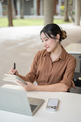 Asian woman student freelancer writing in notebook over laptop sitting at table in open air building