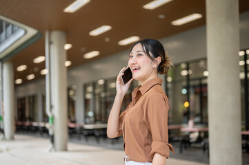 Fototapeta premium Asian woman student talking on smartphone looking ahead while standing sideway in open air building.