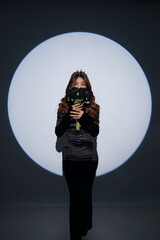 Elegant studio portrait of a glamorous woman wearing a black dress and holding black flowers, photographed against a dramatic circular spotlight background.