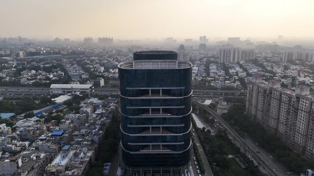 A cinematic aerial drone shot of a modern high-rise commercial tower in Gurugram, India, rising above a dense urban landscape. The camera faces the curved glass facade of the building, revealing