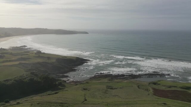 A drone flight reveals the coast of Comillas, Cantabria.The light of the setting sun is increasingly reflected on the sea as the town's beach appears, with a small peninsula visible along the shore.