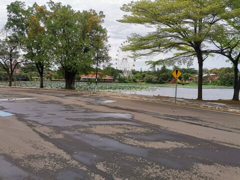 Wet road beside TMII lake, reflective asphalt after rain with calm water and greenery, urban park environment and outdoor transportation scene.