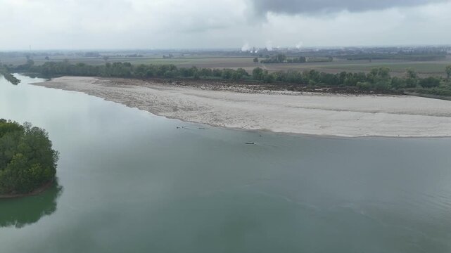 Dynamic river training scene showing a group of Cremona-based rowers gliding across the Po River in coordinated formation, accompanied by a coach shouting instructions from a trailing motorboat