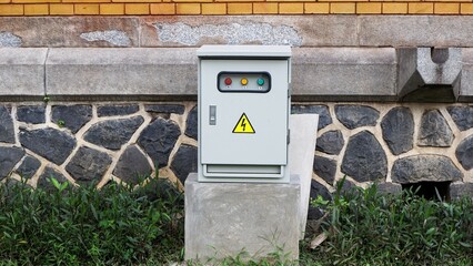 An outdoor electrical control panel with a yellow high voltage warning sign and status lights, mounted on a concrete base against a stone and brick wall in a garden area.