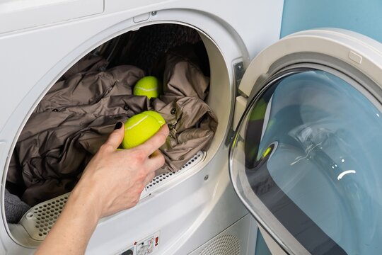 A woman adds tennis balls to a tumble dryer with a down jacket to fluff it while drying. This helps keep the down filling from clumping and maintains its volume for even drying.