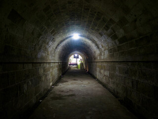 Obraz premium Dark stone tunnel with arched ceiling and light at the exit. Dark arched stone tunnel leading to light. Strong perspective and mystery mood in dark passage.
