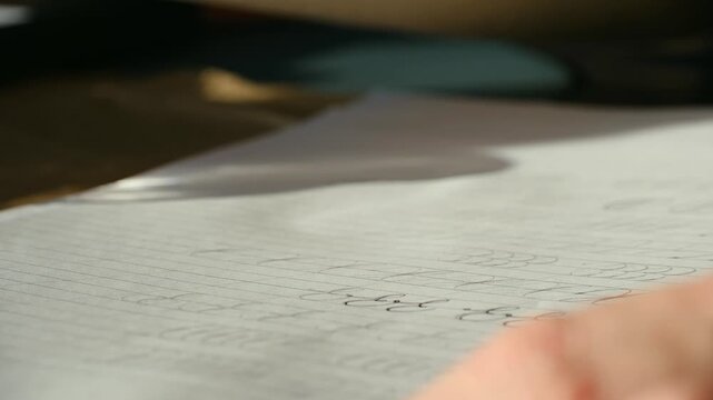 Close up view of a hand practicing calligraphy exercises on lined paper with an old fashioned nib pen, creating elegant cursive letters with black ink under warm, natural sunlight