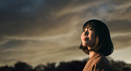 美しい夕焼け・朝焼け　夕日・朝日に照らされた空と日の出の光を浴びた背景　空を見上げ上を向きリラックスした表情の女性の横顔　日本・旅行・観光・アウトドア・リラクゼーションの背景　希望・安堵のイメージ