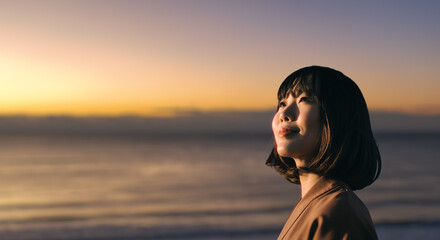 美しい夕焼け・朝焼け　夕日・朝日に照らされた海と日の出の光を浴びた背景　笑顔で空を見上げ上を向きリラックスした表情の女性の横顔　日本・旅行・観光・アウトドア・リラクゼーションの背景　希望・安堵のイメージ　ビジネスはじまり・出勤・転職・就職