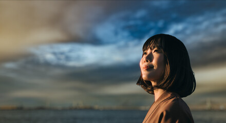 美しい夕焼け・朝焼け　夕日・朝日に照らされた海と日の出の光を浴びた背景　笑顔で空を見上げ上を向きリラックスした表情の女性の横顔　日本・旅行・観光・アウトドア・リラクゼーションの背景　希望・安堵のイメージ　ビジネスはじまり・出勤・転職・就職