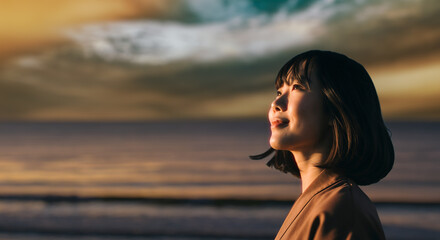 美しい夕焼け・朝焼け　夕日・朝日に照らされた海と日の出の光を浴びた背景　笑顔で空を見上げ上を向きリラックスした表情の女性の横顔　日本・旅行・観光・アウトドア・リラクゼーションの背景　希望・安堵のイメージ　ビジネスはじまり・出勤・転職・就職