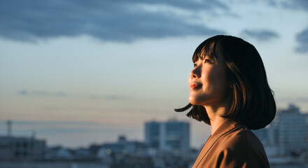 美しい夕焼け・朝焼け　夕日・朝日に照らされた空と街が日の出の光を浴びた背景　空を見上げ上を向きリラックスした表情の女性の横顔　日本・ビジネス・転職出勤・旅行・観光・アウトドア・リラクゼーションの背景　希望・安堵のイメージ