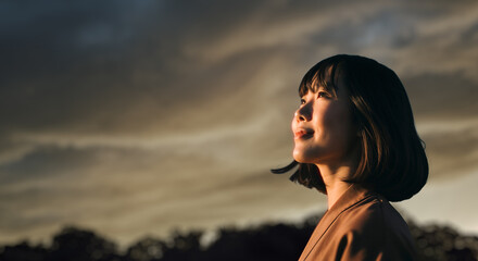 美しい夕焼け・朝焼け　夕日・朝日に照らされた空と日の出の光を浴びた背景　空を見上げ上を向きリラックスした表情の女性の横顔　日本・旅行・観光・アウトドア・リラクゼーションの背景　希望・安堵のイメージ