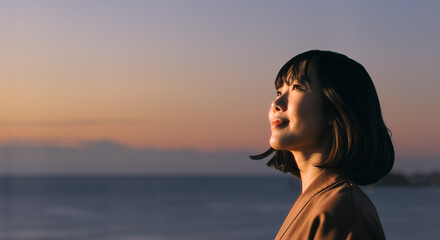 美しい夕焼け・朝焼け　夕日・朝日に照らされた海と日の出の光を浴びた背景　笑顔で空を見上げ上を向きリラックスした表情の女性の横顔　日本・旅行・観光・アウトドア・リラクゼーションの背景　希望・安堵のイメージ　ビジネスはじまり・出勤・転職・就職