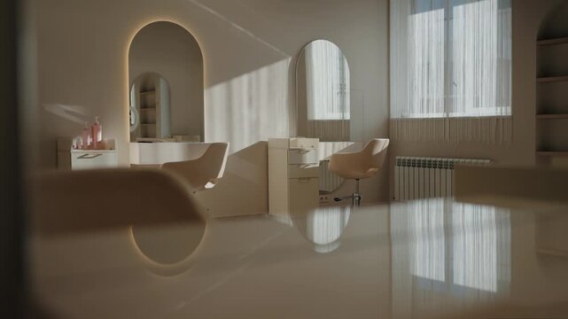 Empty modern hair salon interior with light beige furniture and illuminated arch mirrors. Beauty salon chairs, table and professional tools reflected in glossy surface. Sunlight and shadows in room.